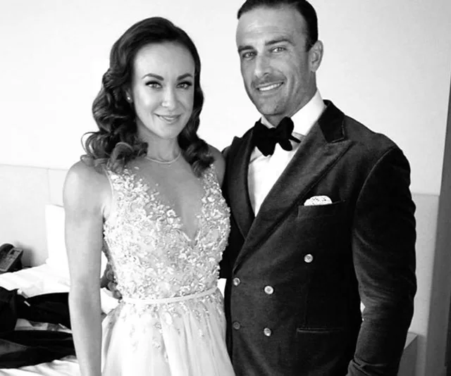 Couple dressed in formal attire, woman in an embellished gown, man in a tuxedo with a bow tie, posing together indoors.