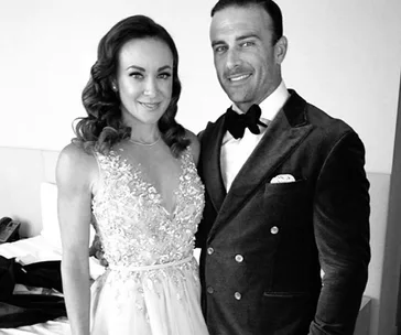 Couple dressed in formal attire, woman in an embellished gown, man in a tuxedo with a bow tie, posing together indoors.
