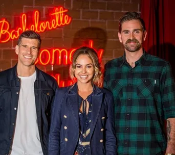 "Three people smiling in front of a 'Bachelorette Comedy' neon sign on a brick wall background."