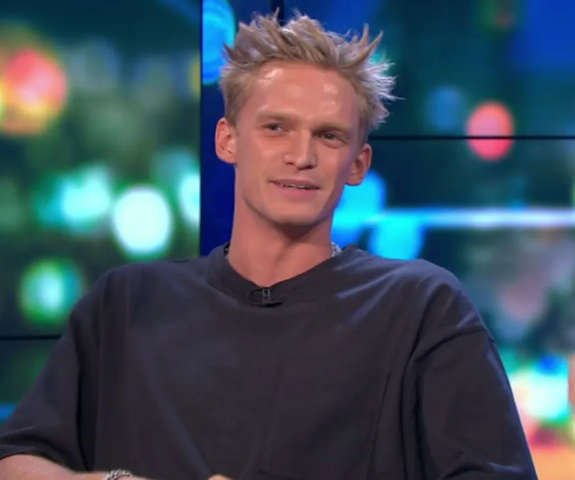 Person with spiky blonde hair and dark shirt smiling on a colorful, blurry background during a talk show appearance.