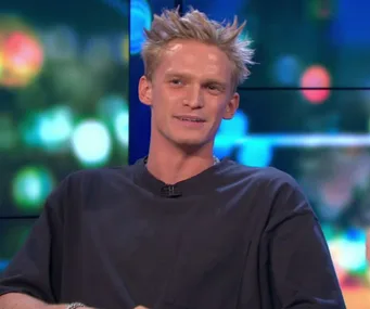 Person with spiky blonde hair and dark shirt smiling on a colorful, blurry background during a talk show appearance.