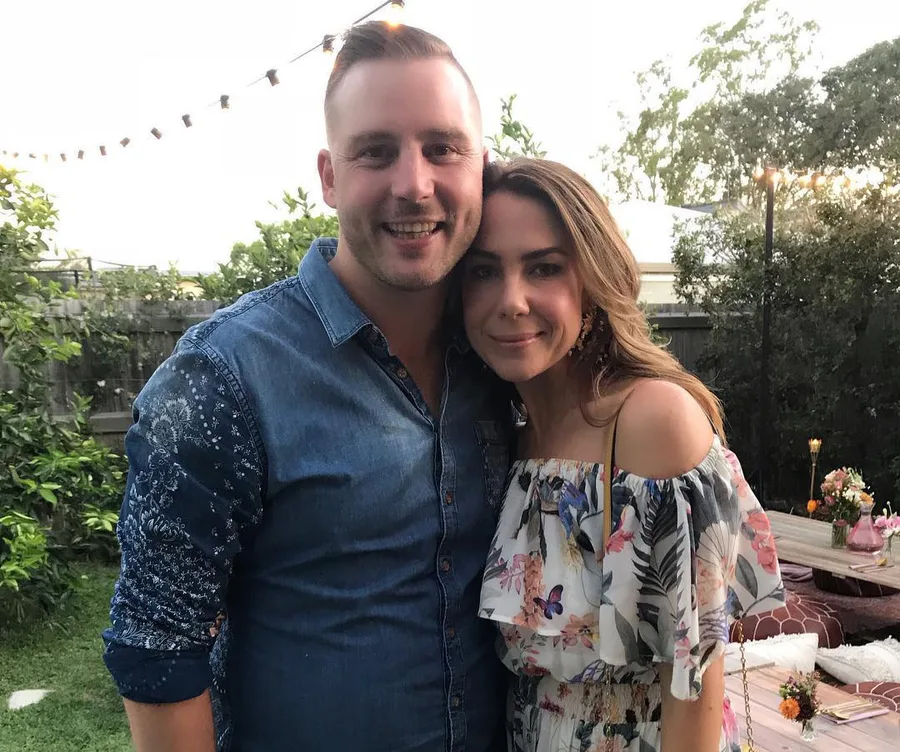 A couple smiling and standing close together at an outdoor gathering, surrounded by greenery and string lights.