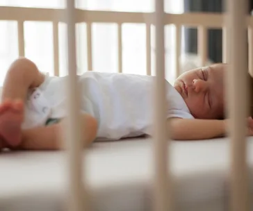 A baby sleeps peacefully in a white crib, wearing a white onesie, with its arms raised above its head.
