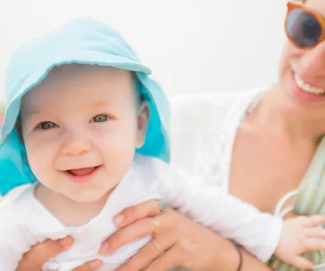 Baby wearing a blue sun hat, smiling, held by a woman wearing sunglasses.