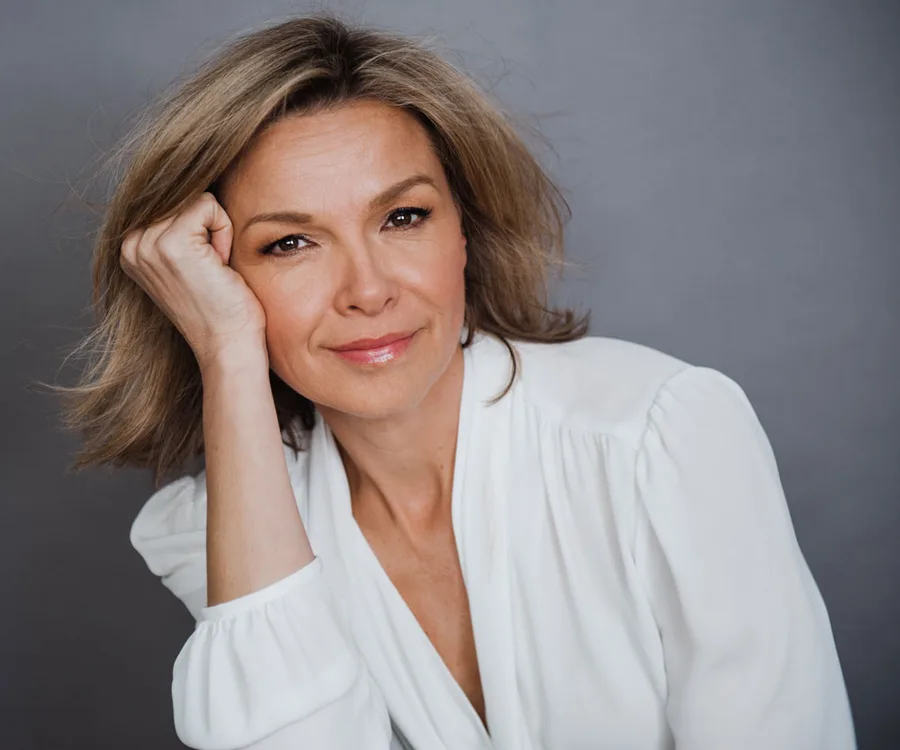 A woman with shoulder-length hair in a white blouse, leaning her head on her hand, smiling gently at the camera.