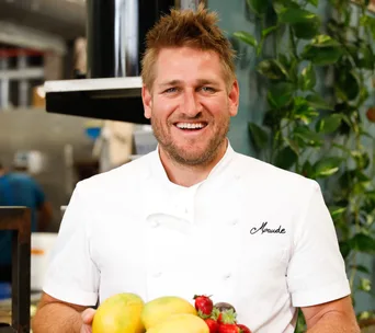 Chef in a white coat, smiling, holding fresh fruits in a kitchen.