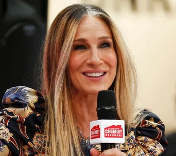 A woman with long hair speaks into a microphone with a "Chemist Warehouse" logo, smiling warmly.