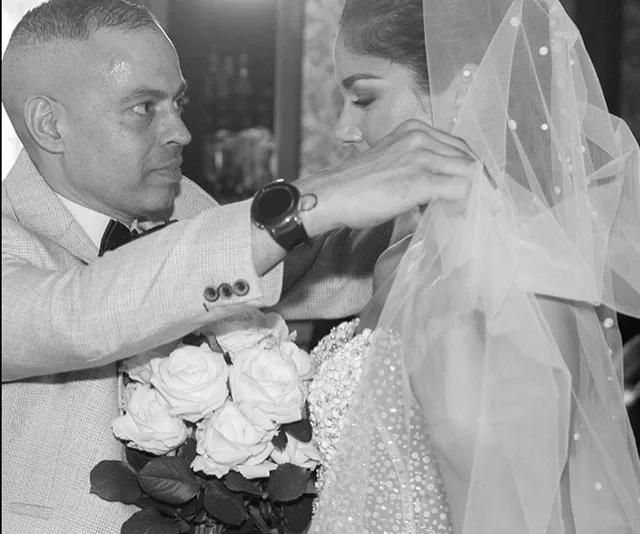 A man helps a bride with her veil; she holds a bouquet of roses. Black and white wedding photo.