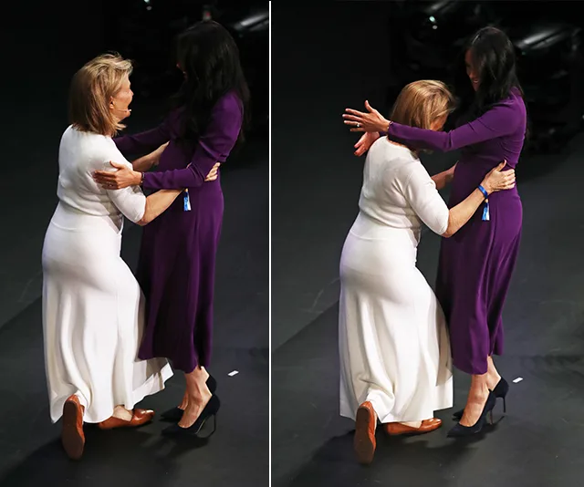 Two women engage in a curtsy and hug onstage, one in a white dress and the other in a purple dress.