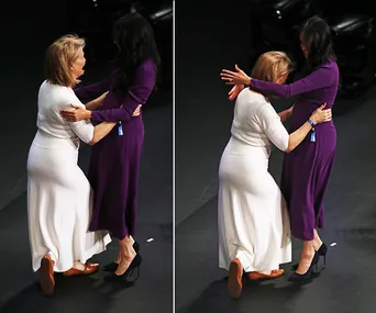 Two women engage in a curtsy and hug onstage, one in a white dress and the other in a purple dress.