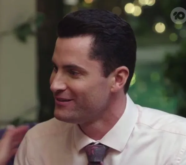 A man in formal attire talking and smiling in a softly lit setting, with blurred greenery in the background.