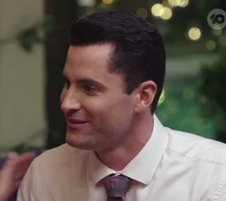 A man in formal attire talking and smiling in a softly lit setting, with blurred greenery in the background.