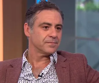 A man in a patterned shirt and blazer appears in a close-up portrait with a neutral expression on a TV set.