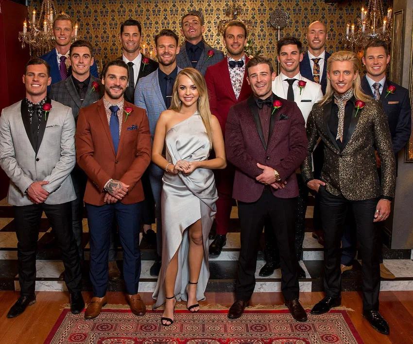 Group photo of a woman in a silver dress surrounded by 13 men in suits, each wearing a rose boutonniere.