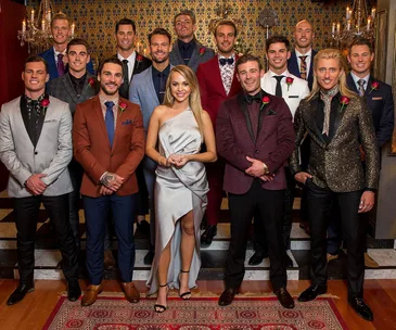 Group photo of a woman in a silver dress surrounded by 13 men in suits, each wearing a rose boutonniere.