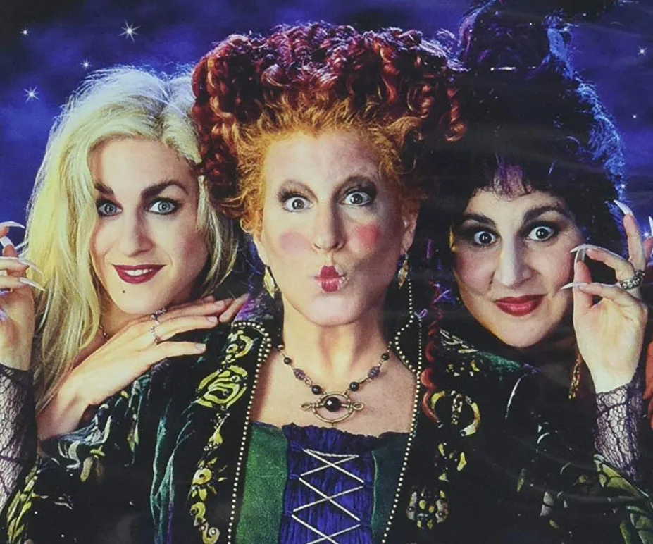 Three women dressed as witches with colorful hair and outfits, posing playfully against a starry backdrop.