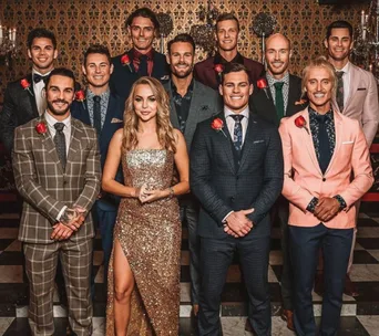 A woman in a glittering dress stands with 11 men in suits wearing red roses on their jackets, set in a formal room.