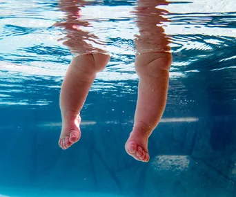 Baby legs underwater in a pool, creating ripples on the surface above.