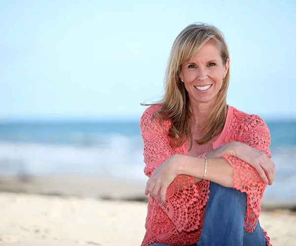 A woman in a coral lace blouse smiles while sitting on a beach with ocean waves in the background.