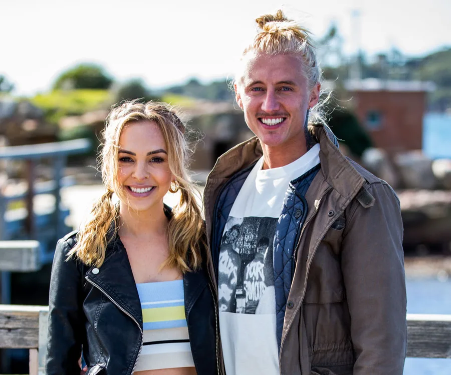 A smiling couple poses together outdoors on a sunny day, with a scenic background.