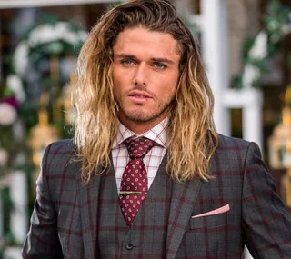A man with long hair in a plaid suit and tie stands outdoors.
