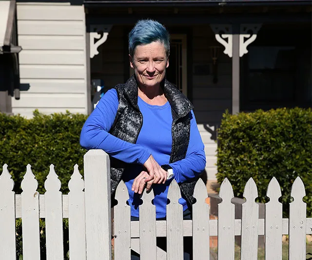 A person with blue hair and a blue top stands smiling by a white picket fence, with a house in the background.
