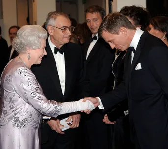 Queen Elizabeth II shakes hands with an actor dressed in a tuxedo at a formal event.