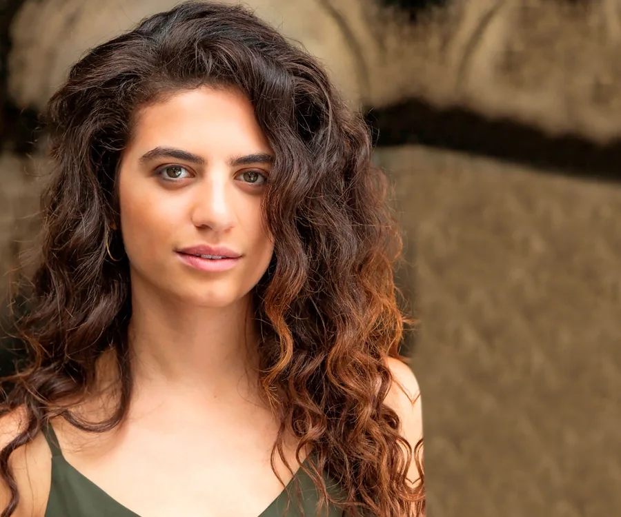 A woman with curly brown hair and a green top stands against a blurred background.