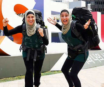 Two women in hijabs smiling and cheering with backpacks during a race, posing in front of a large outdoor sculpture.