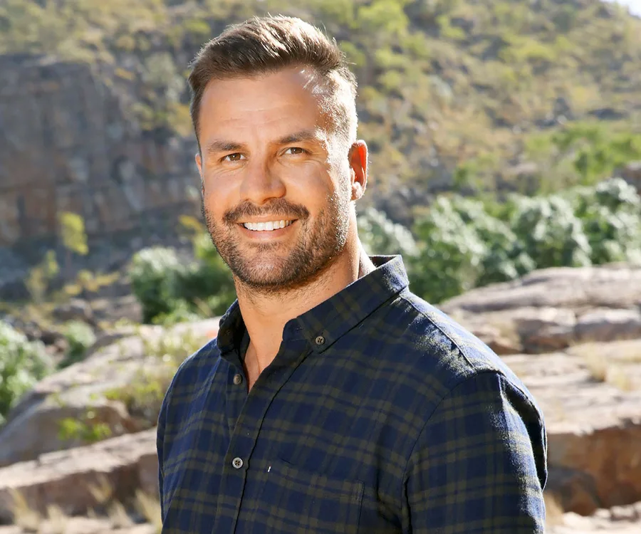 Smiling man in a checkered shirt standing outdoors in a scenic, rocky landscape.