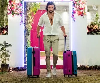 Man in white shirt and beige pants carrying two colorful suitcases, walking out of a lit doorway with flowers.