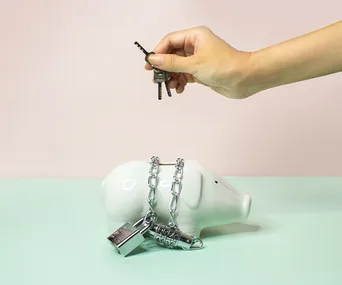 Hand holding keys over a chained and padlocked piggy bank, symbolizing financial restriction or control.
