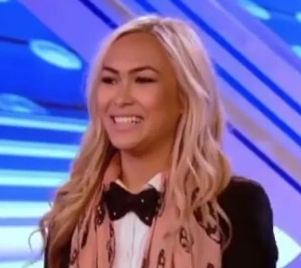 Smiling woman with long blonde hair on a talent show stage, wearing a peach scarf and black bow-tie.