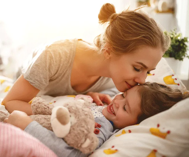 Mother and child cuddling in bed; the child holds a teddy bear and smiles brightly.