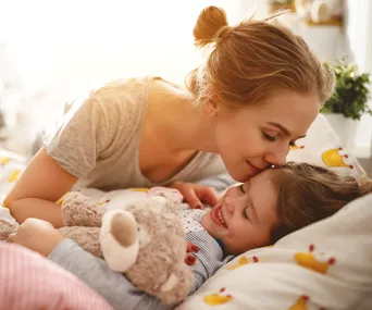 Mother and child cuddling in bed; the child holds a teddy bear and smiles brightly.
