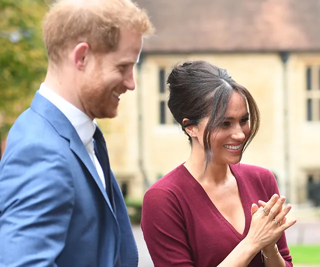 A smiling couple stands outdoors, one in a blue suit and the other in a burgundy outfit, in a sunny setting.