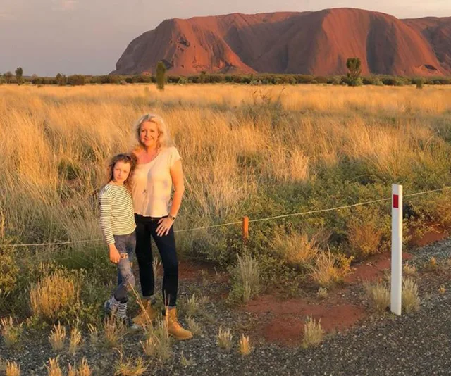 Woman and child standing in front of Uluru at sunset, surrounded by grassland.