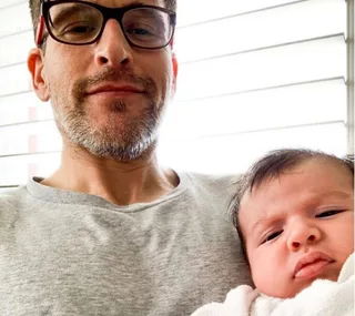 Man wearing glasses holding a baby, both looking at the camera, with light coming through blinds in the background.