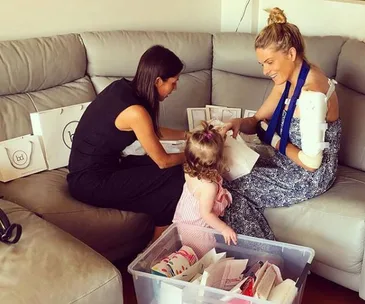 Two women and a child sitting on a couch, with shopping bags and a storage box. One woman has a cast on her arm.