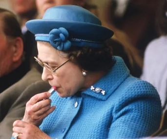A woman in a blue outfit and hat applies lipstick, sitting among an audience.