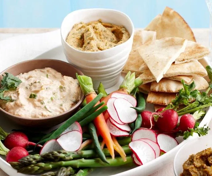 A platter with vegetable sticks, radishes, pita bread, and two bowls of dip, likely suitable for a party or gathering.