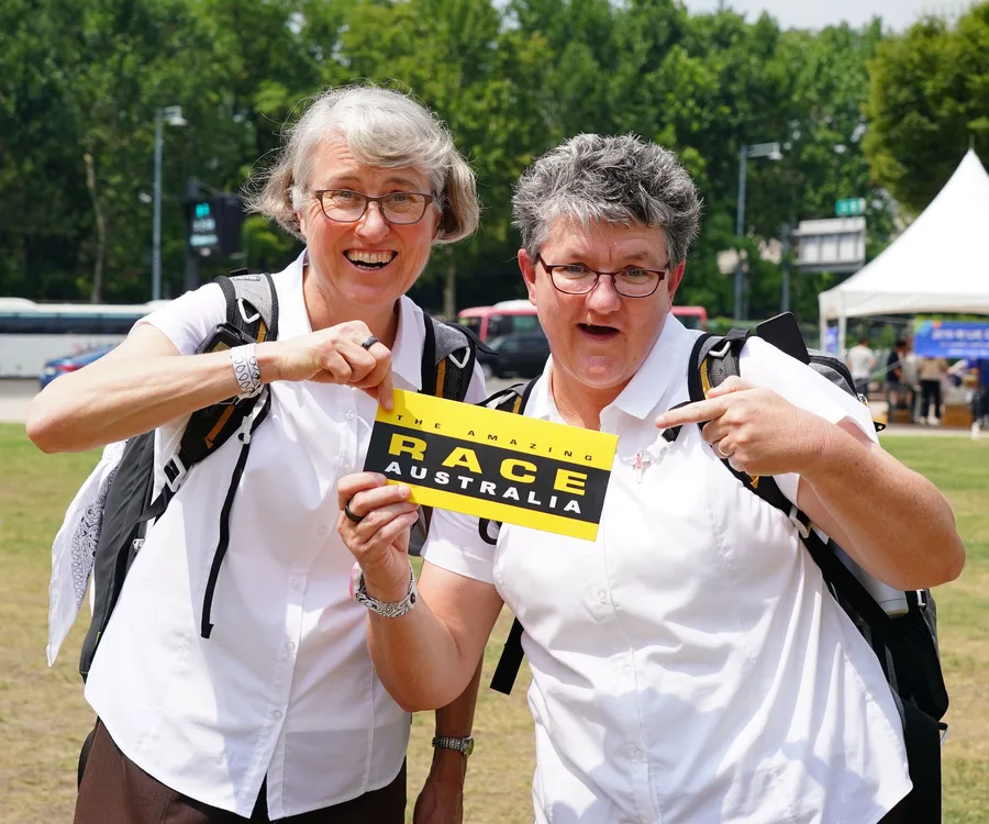 Two women with backpacks hold an "Amazing Race Australia" sign outdoors, smiling excitedly.