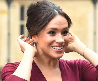 A person smiling, wearing a burgundy top, adjusting earrings with hands near face, hair styled up.