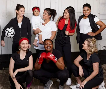 Group of women in sports outfits, one holding a baby, smiling and posing confidently together.