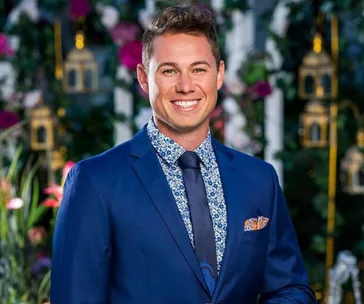 Man in a blue suit with a floral shirt smiling, standing in front of a garden backdrop with lanterns.
