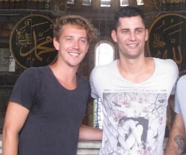 Two men posing and smiling in front of a decorated interior with Arabic script.