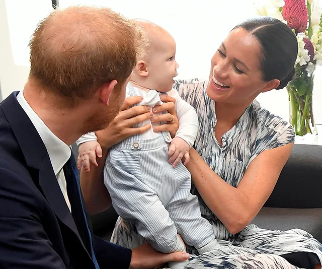 Royal couple with their baby smiling, seated close together in a cozy setting.