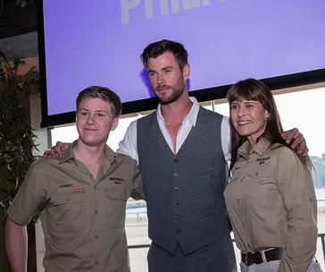 Three people standing together, smiling, with an indoor background and a screen displaying text in the back.