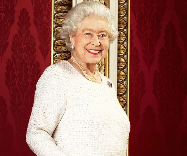 Queen Elizabeth II smiling, wearing a white outfit with pearls, against an ornate red and gold backdrop.