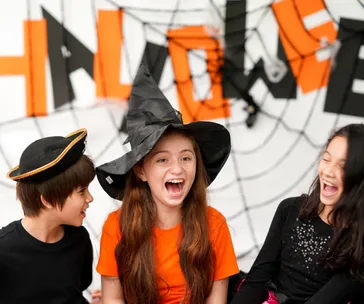 Three kids in costumes laughing at a Halloween party, with festive decorations in the background.